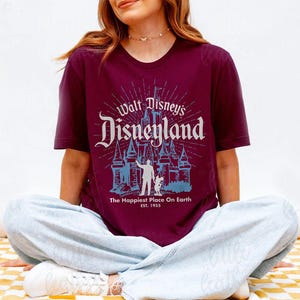 Puede incluir: Camiseta burdeos con el texto "Walt Disney's Disneyland" en un estilo vintage. El dise&ntilde;o incluye un castillo y la frase "The Happiest Place On Earth" con el a&ntilde;o 1955.