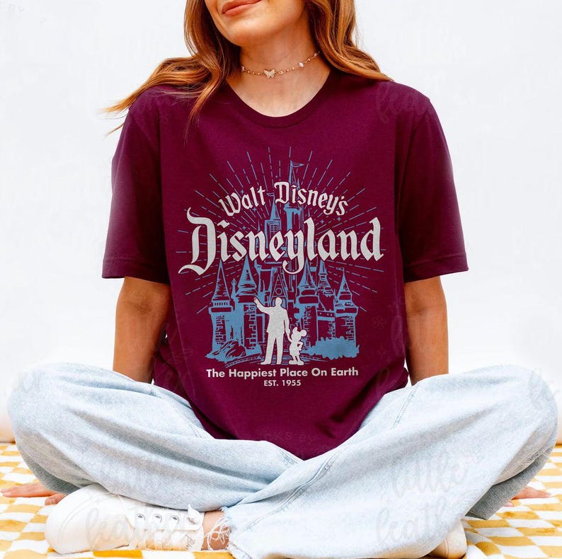 Puede incluir: Camiseta burdeos con el texto "Walt Disney's Disneyland" en un estilo vintage. El dise&ntilde;o incluye un castillo y la frase "The Happiest Place On Earth" con el a&ntilde;o 1955.