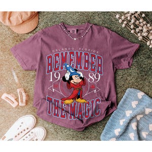 K&ouml;nnte beinhalten: Burgunderfarbenes T-Shirt mit einer Grafik von Mickey Mouse in einem Zaubererkost&uuml;m und dem Text "REMEMBER THE MAGIC" und "ORLANDO, FLORIDA 1989". Das Shirt ist mit einer Halskette, Schuhen und einer blauen Decke gestylt.