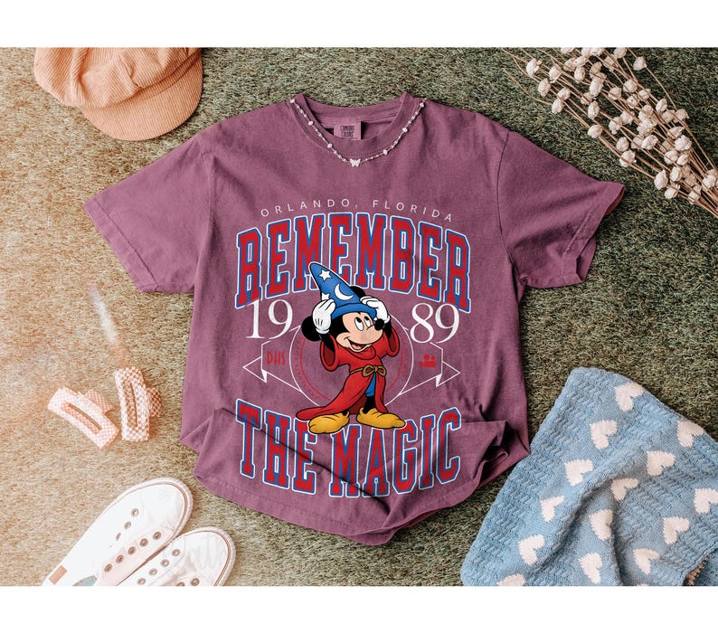 K&ouml;nnte beinhalten: Burgunderfarbenes T-Shirt mit einer Grafik von Mickey Mouse in einem Zaubererkost&uuml;m und dem Text "REMEMBER THE MAGIC" und "ORLANDO, FLORIDA 1989". Das Shirt ist mit einer Halskette, Schuhen und einer blauen Decke gestylt.