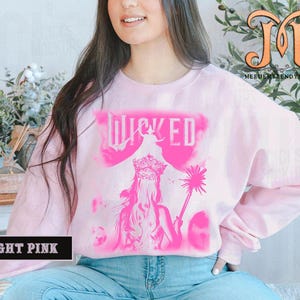 May include: Light pink sweatshirt with a pink graphic of the word "WICKED" and a silhouette of a woman wearing a crown and holding a wand. The sweatshirt is a long-sleeved crew neck.