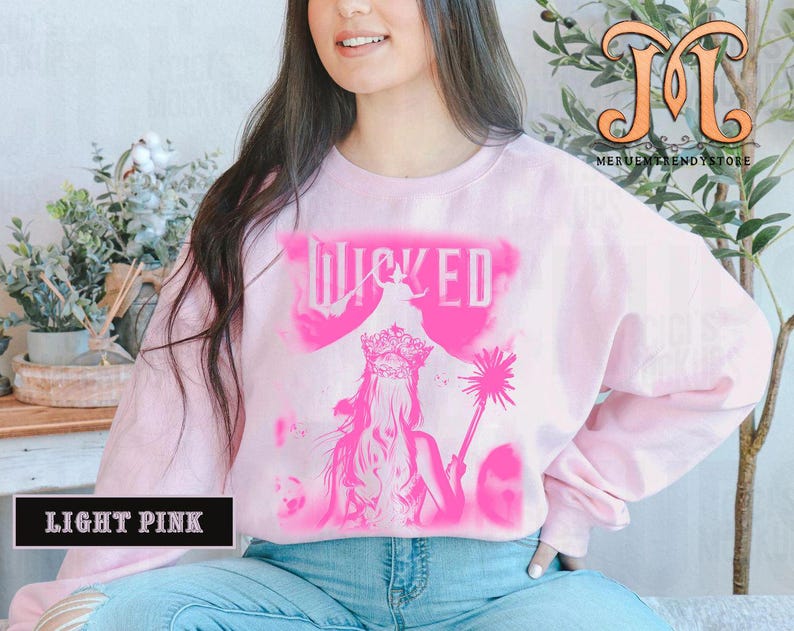 May include: Light pink sweatshirt with a pink graphic of the word "WICKED" and a silhouette of a woman wearing a crown and holding a wand. The sweatshirt is a long-sleeved crew neck.