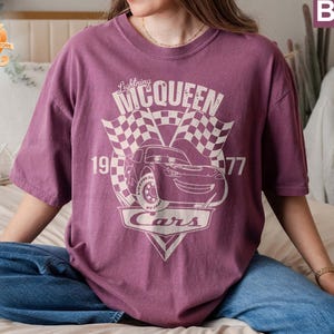 May include: A purple t-shirt with a graphic of Lightning McQueen from the movie Cars. The graphic includes the text "Lightning McQueen", "1997", and "Cars".