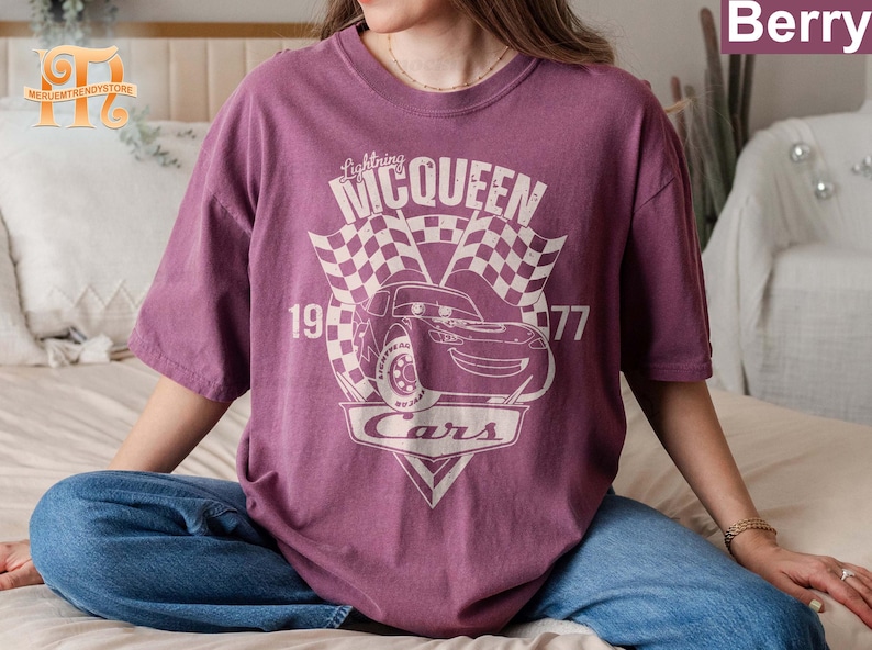May include: A purple t-shirt with a graphic of Lightning McQueen from the movie Cars. The graphic includes the text "Lightning McQueen", "1997", and "Cars".