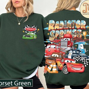 Puede incluir: Sudadera verde oscuro con un dise&ntilde;o de coche de carreras verde y la palabra "name" en la parte delantera. La parte trasera presenta un dise&ntilde;o colorido de personajes y las palabras "Radiator Springs Cars Lightning McQueen". El texto "Forset Green" est&aacute; en la parte inferior.