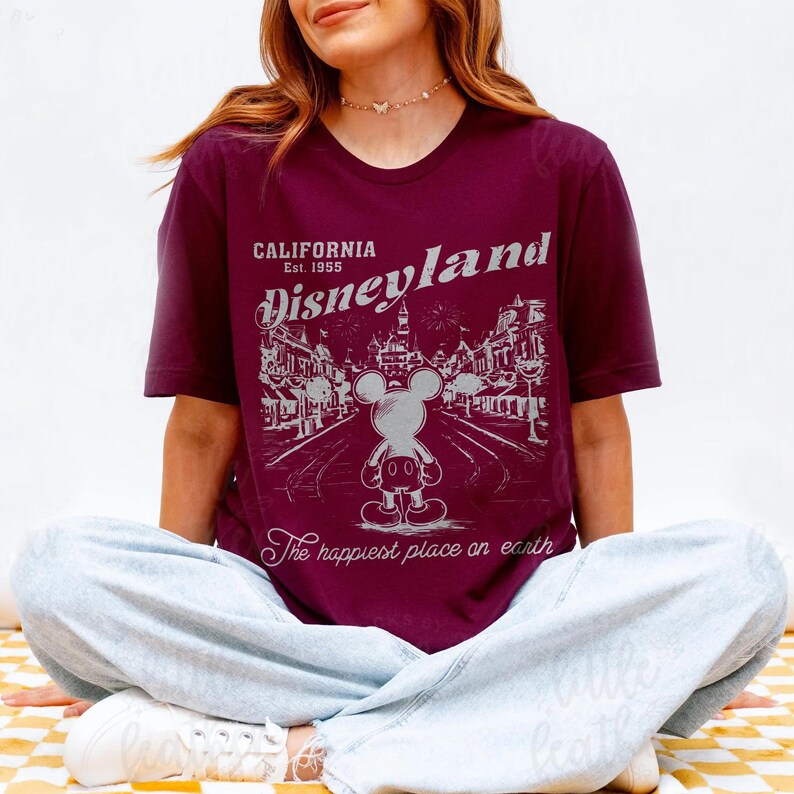 以下が含まれることがあります： ディズニーランドのグラフィックがプリントされたバーガンディ色のTシャツ。デザインにはミッキーマウス、「California Est. 1955 Disneyland」と「The happiest place on earth」の文字が描かれています。Tシャツは、あぐらをかいて座っている人が着用しています。