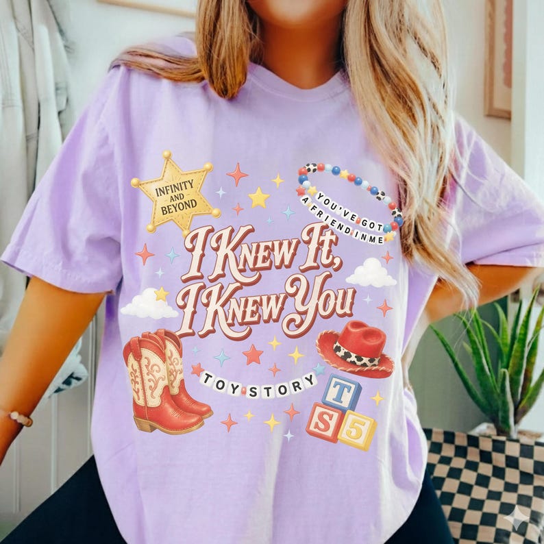 May include: A light purple t-shirt featuring a graphic print with the text "I Knew It, I Knew You" and "Toy Story". The design includes a yellow sheriff star, a beaded bracelet, a red cowboy hat, cowboy boots, and alphabet blocks.