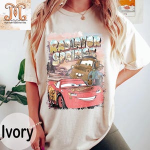 May include: Ivory t-shirt with a vintage-style graphic of Lightning McQueen and Mater from Cars. The graphic says "Radiator Springs" in a retro font.