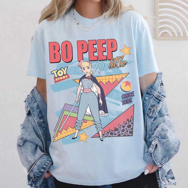 May include: Light blue t-shirt featuring a graphic of Bo Peep from Toy Story. The design includes the text "BO PEEP" in red, "TOY STORY" in yellow, and "SINCE 1995". The graphic also features Bo Peep in a blue outfit with a cape and staff.