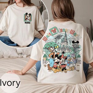 May include: Ivory t-shirt with a Mickey Mouse graphic and the word "Daddy" on the front. The back features a colourful "Walt Disney World" design with Disney characters and a castle.