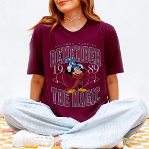 K&ouml;nnte beinhalten: Burgunderfarbenes T-Shirt mit einer Grafik von Mickey Mouse als Zauberer und dem Text "REMEMBER THE MAGIC 1989 ORLANDO, FLORIDA" in Blau und Wei&szlig;. Retro-Design.