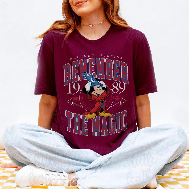K&ouml;nnte beinhalten: Burgunderfarbenes T-Shirt mit einer Grafik von Mickey Mouse als Zauberer und dem Text "REMEMBER THE MAGIC 1989 ORLANDO, FLORIDA" in Blau und Wei&szlig;. Retro-Design.