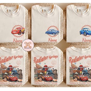 Op de afbeelding: Cr&egrave;mekleurige t-shirts met cartoon autographics. Sommige shirts hebben een enkel auto-ontwerp met het naamveld, terwijl andere een grotere afbeelding tonen met de tekst "Radiator Springs" en "Welcome to Radiator Springs".
