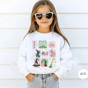 May include: A white sweatshirt featuring a colorful, whimsical design with witch-themed illustrations. The design includes a witch hat, a castle, a broom, and the word "Wicked". The sweatshirt is worn by a young person wearing heart-shaped sunglasses.
