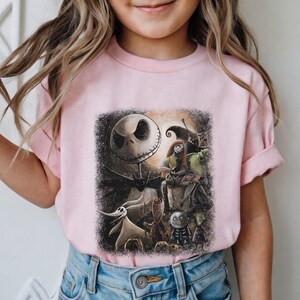 May include: A light pink t-shirt featuring a graphic from "The Nightmare Before Christmas." The design includes Jack Skellington, Sally, Zero, and other characters against a dark, moonlit background. The shirt is made of a soft material.