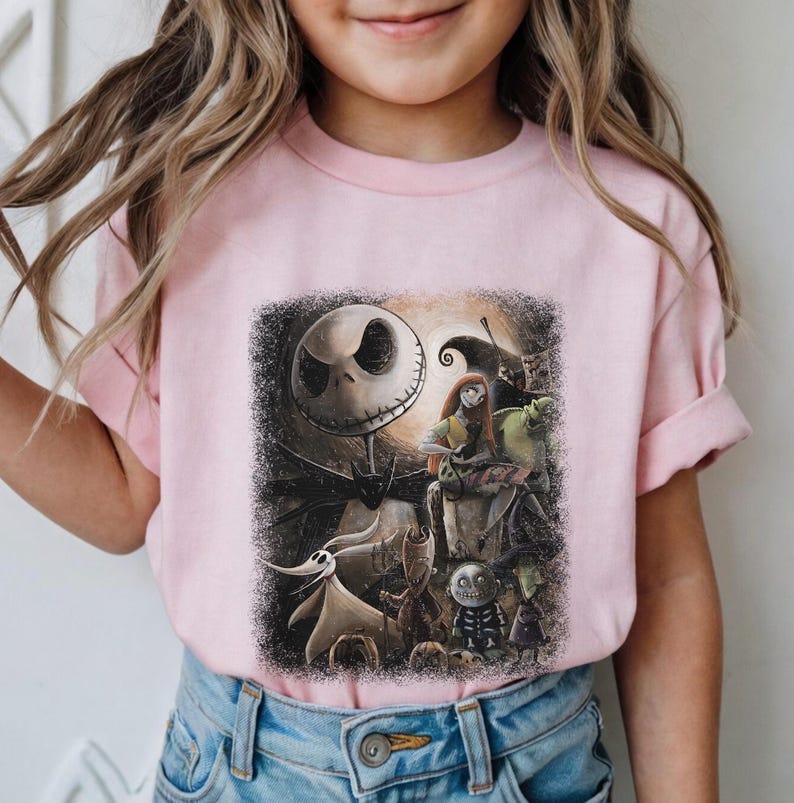 May include: A light pink t-shirt featuring a graphic from "The Nightmare Before Christmas." The design includes Jack Skellington, Sally, Zero, and other characters against a dark, moonlit background. The shirt is made of a soft material.
