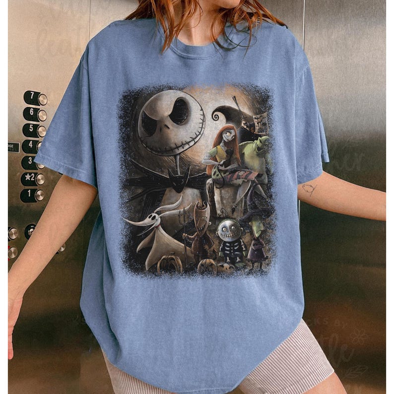 May include: A light blue t-shirt featuring a graphic of characters from The Nightmare Before Christmas. The design includes Jack Skellington, Sally, Zero, and other characters in a dark, detailed illustration. The shirt has short sleeves and a crew neck.