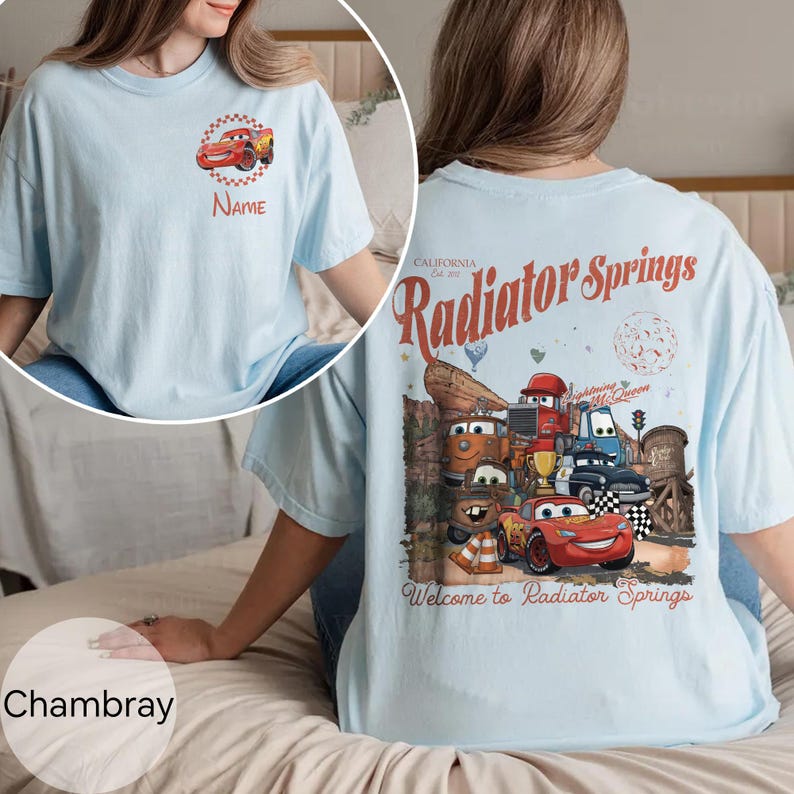 Op de afbeelding: Lichtblauw T-shirt met een cartoon auto-ontwerp en het woord "Name" op de voorkant. De achterkant van het shirt toont een afbeelding van cartoonauto's met de woorden "Radiator Springs" en "Welcome to Radiator Springs".