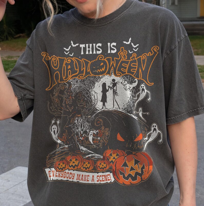 May include: A charcoal gray short-sleeve t-shirt featuring a graphic print with the text "THIS IS HALLOWEEN" and "EVERYBODY MAKE A SCENE!" The design includes silhouettes of two figures, ghosts, a large pumpkin, and smaller pumpkins in orange and white.