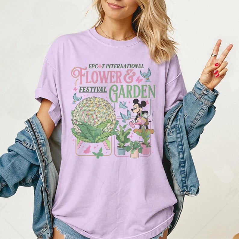 May include: A light purple t-shirt featuring the text "EPCOT INTERNATIONAL FLOWER & FESTIVAL GARDEN" with a graphic of Mickey Mouse, butterflies, and a large floral sphere. The shirt has short sleeves.