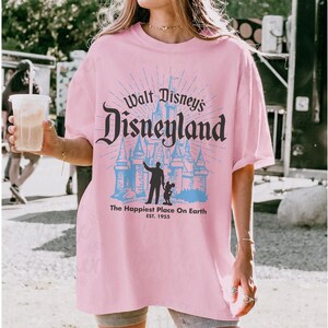 Puede incluir: Camiseta rosa claro con el texto "Walt Disney's Disneyland" en un estilo vintage. El dise&ntilde;o incluye la silueta de un castillo y la frase "The Happiest Place On Earth" con el a&ntilde;o de establecimiento, 1955.