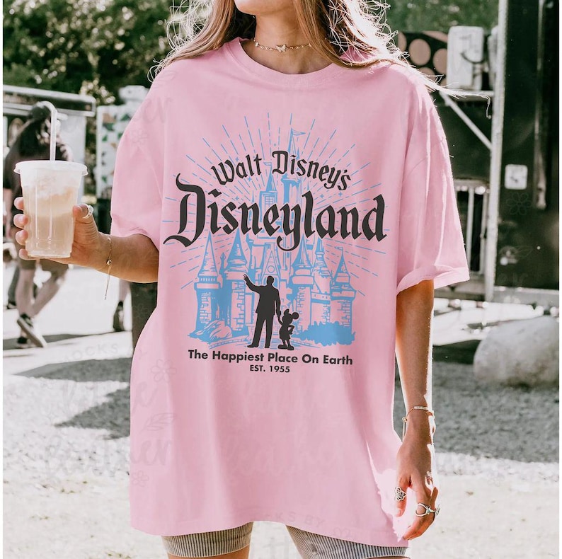 Puede incluir: Camiseta rosa claro con el texto "Walt Disney's Disneyland" en un estilo vintage. El dise&ntilde;o incluye la silueta de un castillo y la frase "The Happiest Place On Earth" con el a&ntilde;o de establecimiento, 1955.