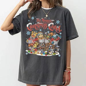 Puede incluir: Camiseta gris oscuro con un gráfico de los personajes de los Muppets de "The Muppet's Christmas Carol". El diseño incluye el título en rojo y blanco, con copos de nieve. Los Muppets están representados cantando villancicos. La camiseta es de corte holgado.
