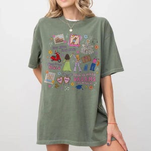 May include: An olive green t-shirt featuring a vibrant graphic. The design incorporates illustrations of a scooter, figures, and the text "THIS IS WHAT DREAMS ARE MADE OF". The t-shirt has short sleeves and a loose fit.