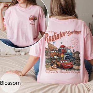 Op de afbeelding: Roze T-shirt met een cartoon auto-ontwerp op de voorkant en een grotere afbeelding op de achterkant met de tekst "Radiator Springs" en "Welcome to Radiator Springs". Het shirt heeft een ronde hals en korte mouwen.