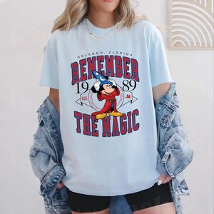 K&ouml;nnte beinhalten: Hellblaues T-Shirt mit einer Grafik von Mickey Mouse in einem Zauberer-Outfit, mit dem Text "REMEMBER THE MAGIC" und "ORLANDO, FLORIDA 1989". Das Design ist in Rot, Wei&szlig; und Schwarz gehalten.
