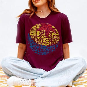 May include: Burgundy crew neck t-shirt featuring a circular graphic design. The graphic is divided into three sections: a red starburst at the top, yellow shapes in the middle, and blue shapes at the bottom. The shirt is worn by a person sitting cross-legged.