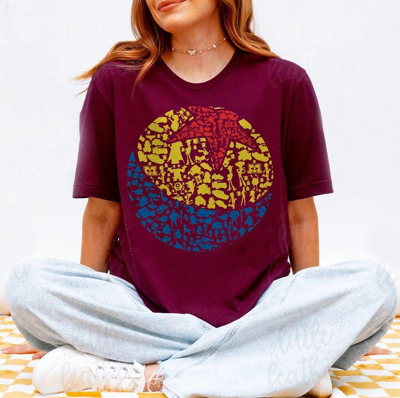 May include: Burgundy crew neck t-shirt featuring a circular graphic design. The graphic is divided into three sections: a red starburst at the top, yellow shapes in the middle, and blue shapes at the bottom. The shirt is worn by a person sitting cross-legged.