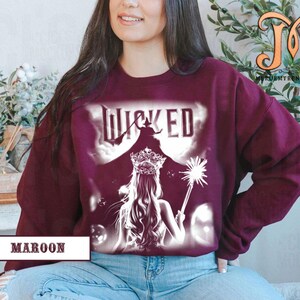 May include: Maroon sweatshirt featuring a graphic from the musical "Wicked." The design includes the word "Wicked" in white, a silhouette of a witch on a broom, and a princess with a crown and wand. The word "Maroon" is printed at the bottom.
