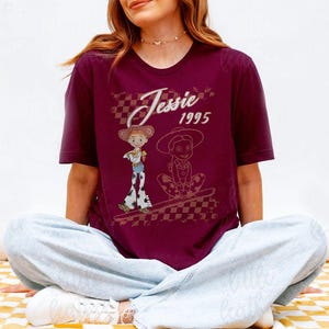 K&ouml;nnte beinhalten: Burgunderfarbenes T-Shirt mit einem Aufdruck der Figur Jessie aus Toy Story und dem Text "Jessie 1995". Das Design zeigt zwei Illustrationen von Jessie, eine stehend und eine sitzend, mit einem karierten Hintergrund.