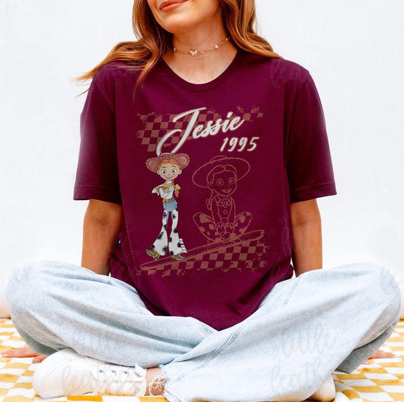 K&ouml;nnte beinhalten: Burgunderfarbenes T-Shirt mit einem Aufdruck der Figur Jessie aus Toy Story und dem Text "Jessie 1995". Das Design zeigt zwei Illustrationen von Jessie, eine stehend und eine sitzend, mit einem karierten Hintergrund.