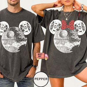 May include: Two dark gray t-shirts with a Death Star design, incorporating Mickey Mouse ears. One shirt has a red bow. The Death Star is white and gray. The shirts are oversized and worn by two people.