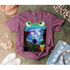 May include: A purple t-shirt with a graphic from the Disney Pixar film Toy Story 5. The design features Woody, Buzz Lightyear, and Jessie. The graphic is framed by a green frog-like border.