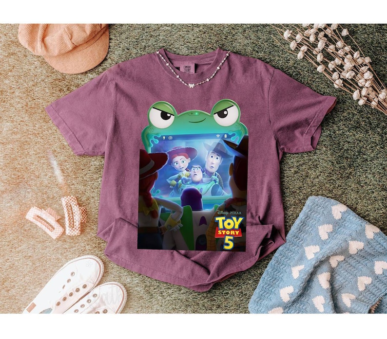 May include: A purple t-shirt with a graphic from the Disney Pixar film Toy Story 5. The design features Woody, Buzz Lightyear, and Jessie. The graphic is framed by a green frog-like border.