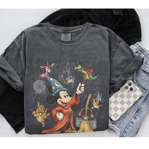 K&ouml;nnte beinhalten: Dunkelgraues Langarmshirt mit einer Grafik von Mickey Mouse als Zauberlehrling, zusammen mit anderen animierten Charakteren und einem Schloss. Das Shirt wird mit Jeans und einer Handyh&uuml;lle gezeigt.