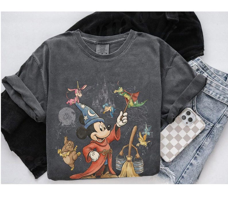 K&ouml;nnte beinhalten: Dunkelgraues Langarmshirt mit einer Grafik von Mickey Mouse als Zauberlehrling, zusammen mit anderen animierten Charakteren und einem Schloss. Das Shirt wird mit Jeans und einer Handyh&uuml;lle gezeigt.
