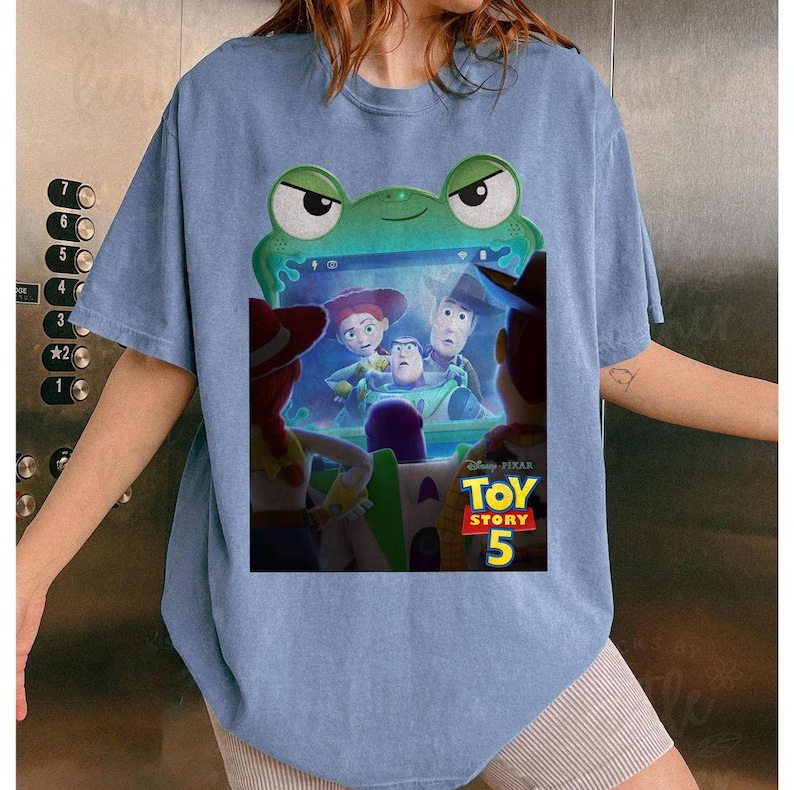 May include: Pale blue t-shirt with a green frog graphic and a screen showing Toy Story 5 characters. The shirt has short sleeves and a loose fit, ideal for everyday wear. The design is a fun, casual style.