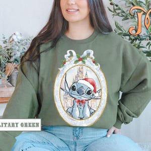 May include: Military green sweatshirt featuring a cartoon character in a Santa hat and bow tie, set against a castle backdrop within a decorative frame. The sweatshirt has the text "MILITARY GREEN" at the bottom.