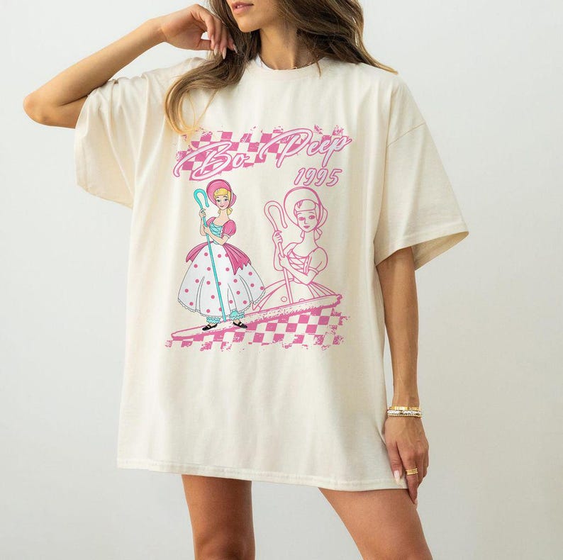 K&ouml;nnte beinhalten: Cremefarbenes T-Shirt mit einem Aufdruck von Bo Peep aus Toy Story. Das Design zeigt zwei Illustrationen von Bo Peep in Rosa und Wei&szlig;, mit dem Text "Bo Peep 1995" in einer rosa, stilisierten Schrift. Das Shirt hat kurze &Auml;rmel und eine lockere Passform.