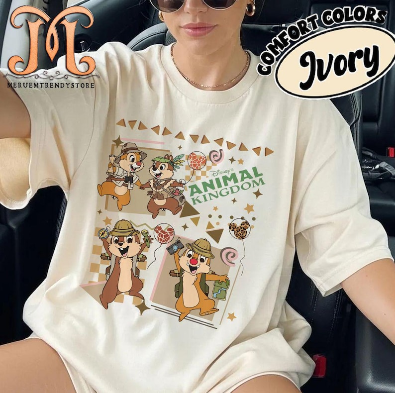 May include: An ivory short-sleeve t-shirt featuring a graphic print of four chipmunks in safari gear. The design includes the text Disney ANIMAL KINGDOM, various balloons, and geometric shapes in brown and tan tones across the front of the garment.