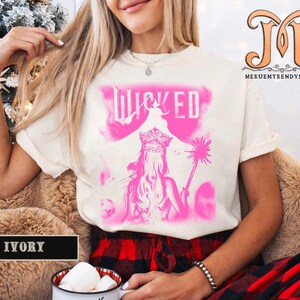 May include: Ivory-colored t-shirt featuring a pink graphic of a woman with a crown and the word "WICKED". The design includes a silhouette of a witch and a magic wand. The shirt is worn by a person.