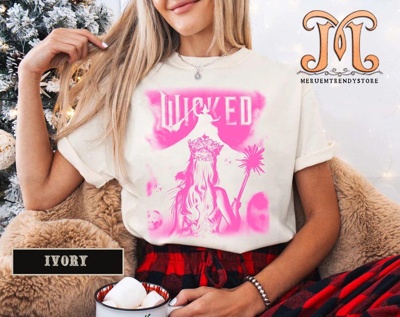 May include: Ivory-colored t-shirt featuring a pink graphic of a woman with a crown and the word "WICKED". The design includes a silhouette of a witch and a magic wand. The shirt is worn by a person.