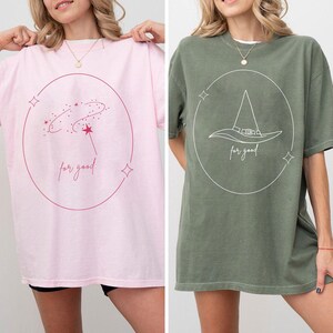Puede incluir: Dos camisetas de gran tamaño, una rosa y otra verde oliva. La camiseta rosa presenta un diseño celestial con las palabras "for good". La camiseta verde muestra un diseño de sombrero de bruja con el mismo texto. Ambas camisetas tienen cuello redondo.