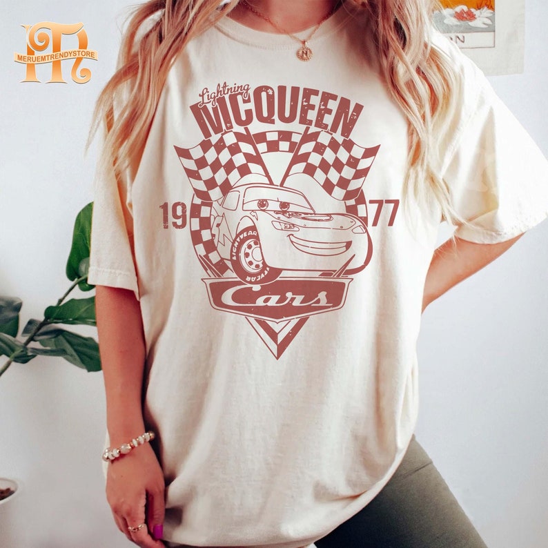May include: A white t-shirt with a red graphic of Lightning McQueen from the Disney Pixar movie Cars. The graphic includes the text "Lightning McQueen", "Cars", and "19-77".