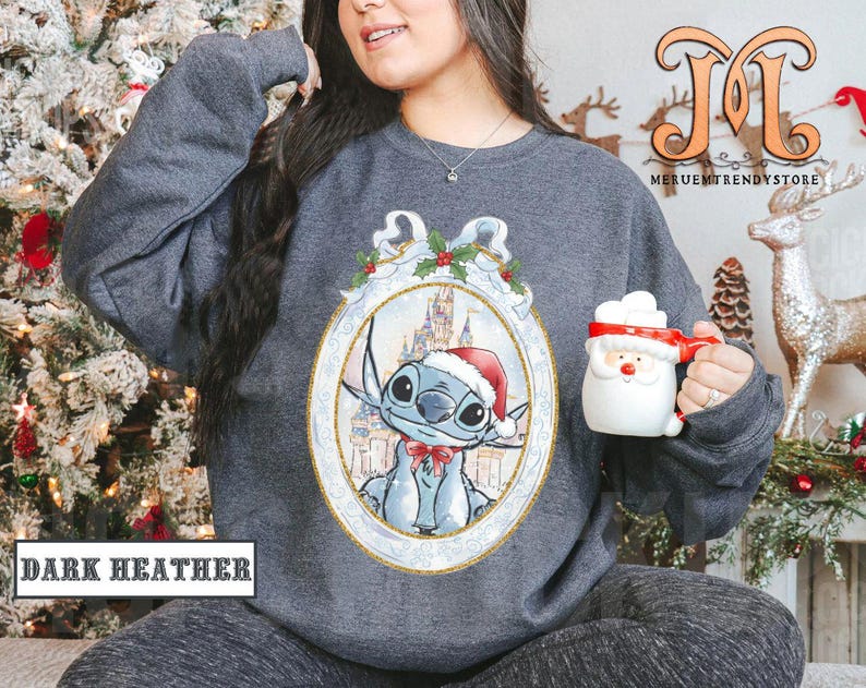May include: Dark heather gray sweatshirt featuring a festive Stitch design in a decorative frame. Stitch is wearing a Santa hat and bow tie. The sweatshirt also has the text "DARK HEATHER". A Santa mug is held in the hand.