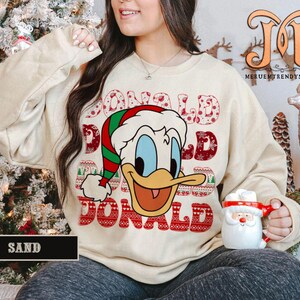 May include: Sand-colored sweatshirt featuring a graphic of Donald Duck in a Santa hat with the name "DONALD" in a festive font. The sweatshirt is part of a Christmas-themed collection.